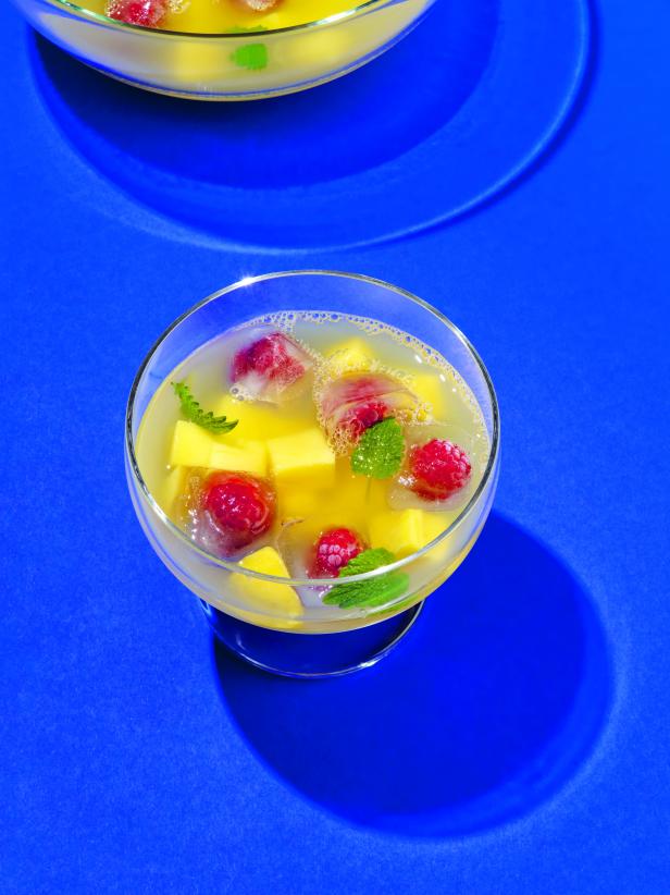 Ein Glas mit einem Getränk, Mangowürfeln, Himbeeren und Minze auf blauem Grund.