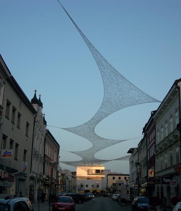 Eine Straße in einer Stadt mit einem ungewöhnlichen Sonnenschutznetz über den Köpfen.