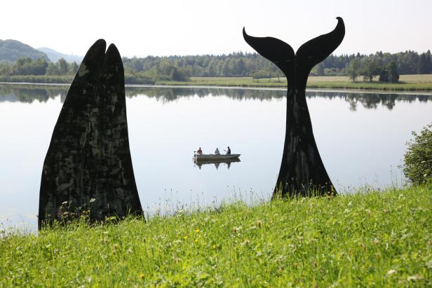 Drei Personen in einem Boot auf einem See, eingerahmt von Walskulpturen.