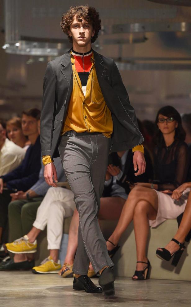 Ein Mann mit lockigem Haar läuft auf einem Laufsteg in einem grauen Anzug und einem gelben Hemd.