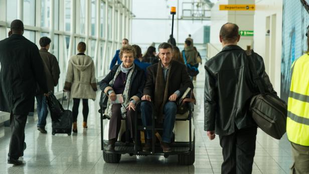 Judi Dench und Steve Coogan sitzen in einem Golfwagen in einem Flughafenterminal.