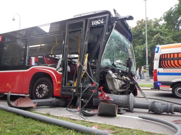Ein roter Bus ist mit einer Straßenlaterne kollidiert und stark beschädigt worden. Ein Rettungswagen steht bereit.