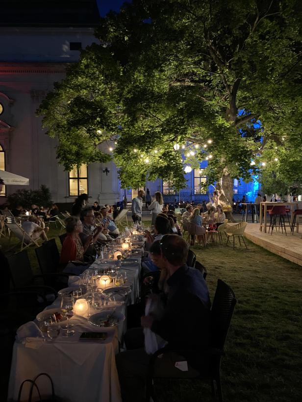 Im Garten eines Restaurants sitzen Menschen an Tischen unter Lichterketten.