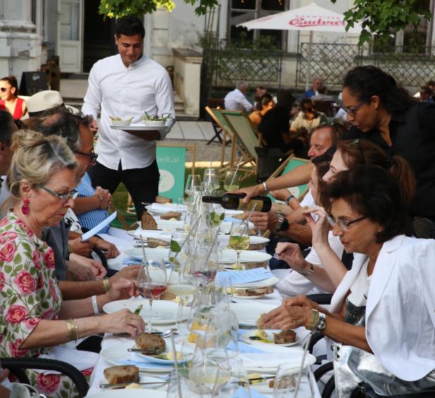 Gäste sitzen an einer langen Tafel im Freien und werden bedient.