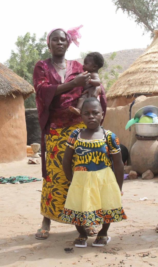 Eine Frau mit einem Baby auf dem Arm und ein Mädchen stehen vor traditionellen Hütten.