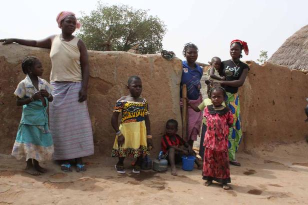 Eine Gruppe von Frauen und Kindern steht vor einer Lehmwand in einem Dorf.