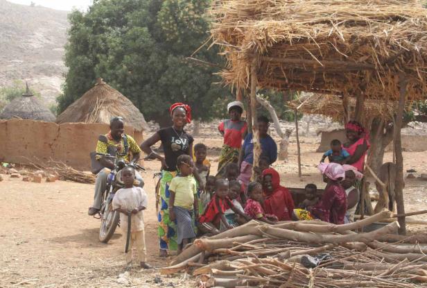 Eine Gruppe von Menschen versammelt sich in einem Dorf unter einem Strohdach.