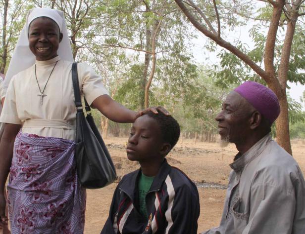 Eine Nonne legt einem Jungen die Hand auf den Kopf, während ein älterer Mann mit lila Mütze daneben sitzt.