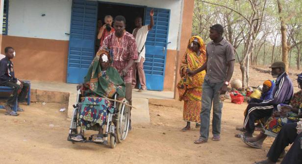 Eine Frau im Rollstuhl und andere Personen mit Augenverbänden stehen vor einem Gebäude.