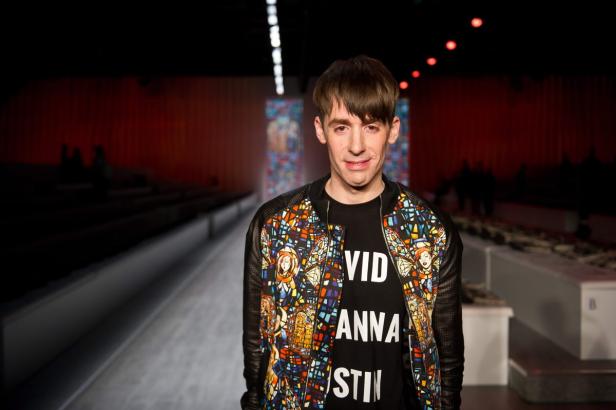 Ein Mann mit einer bunten Jacke und einem T-Shirt mit dem Aufdruck „David, Anna, Kristin“ steht auf einem Laufsteg.