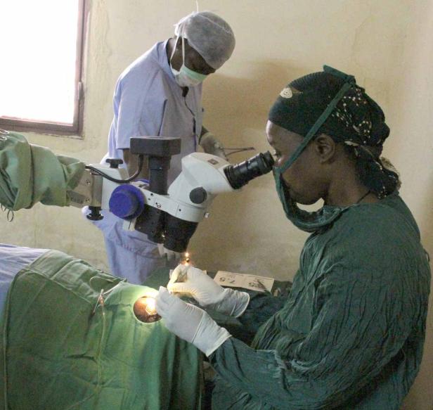Eine Ärztin führt mit einem Mikroskop eine Augenoperation durch, assistiert von einer weiteren Person.