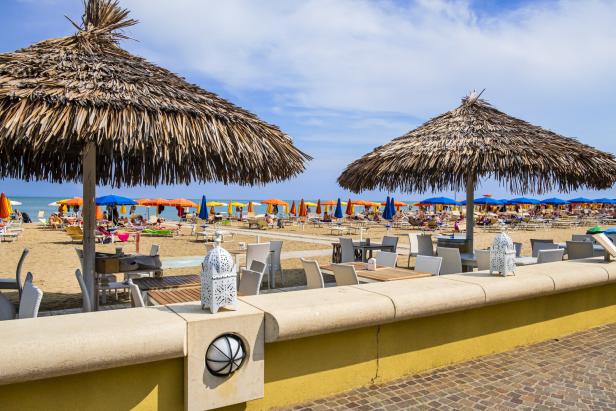 Ein Strand mit bunten Sonnenschirmen und Strohdächern über Tischen und Stühlen.
