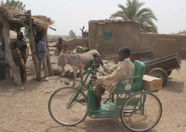 Ein Junge fährt ein grünes Dreirad in einem ländlichen Dorf, während ein Esel daneben steht.