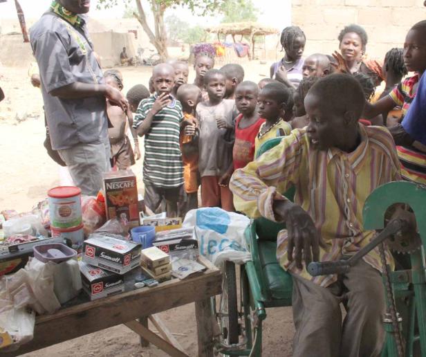 Ein Mann sitzt in einem Rollstuhl vor einem Tisch mit Waren, umgeben von Kindern.