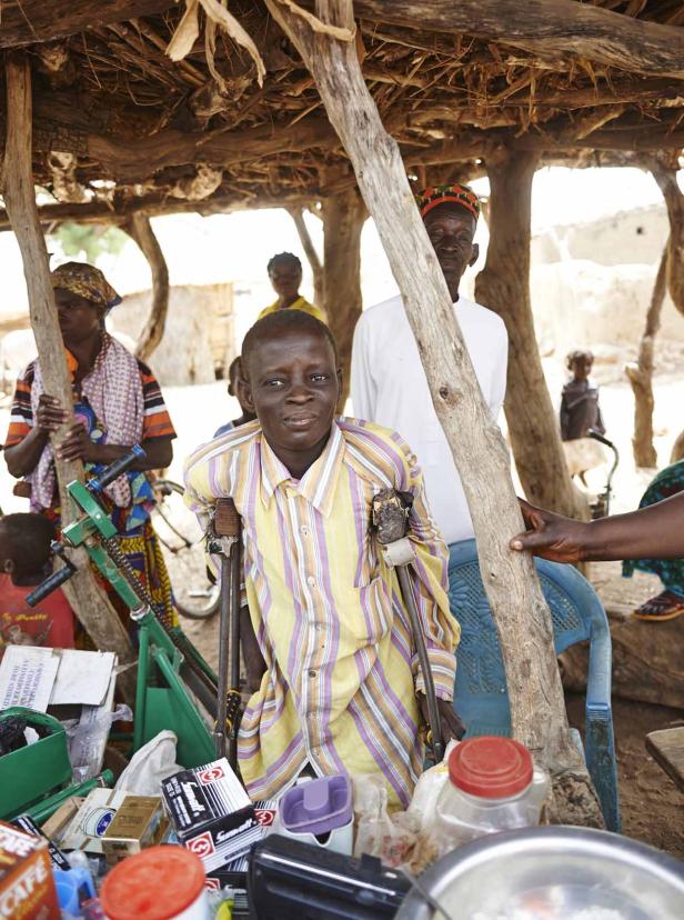 Ein Mann mit Krücken steht unter einem einfachen Unterstand in einem Dorf.