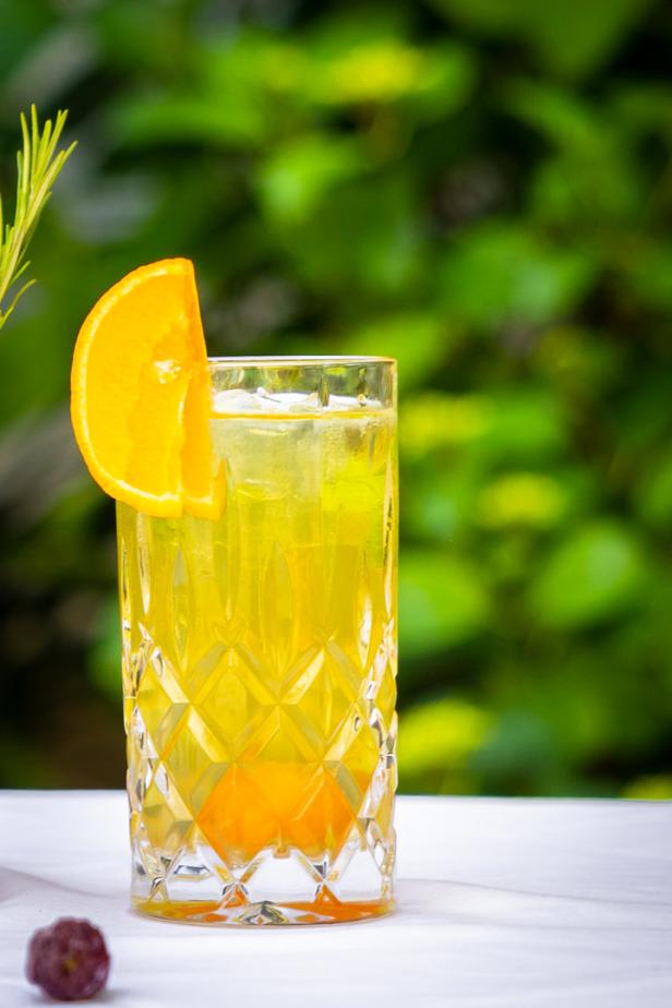 Ein Glas mit einem gelben Getränk, einer Orangenscheibe und Eiswürfeln steht auf einem Tisch.