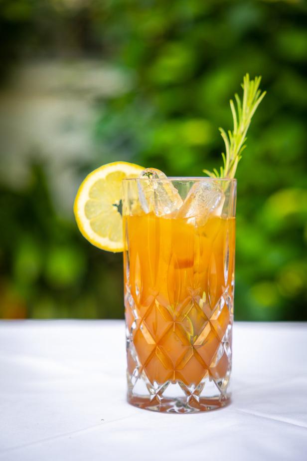 Ein Glas mit einem orangefarbenen Getränk, Eis, einer Zitronenscheibe und einem Rosmarinzweig.