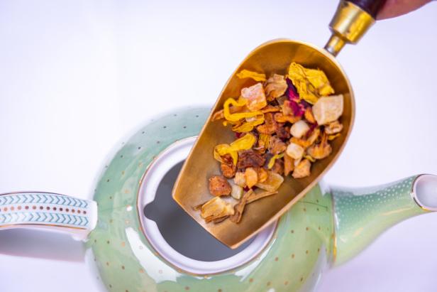 Ein goldener Löffel mit Früchtetee wird in eine hellgrüne Teekanne gefüllt.