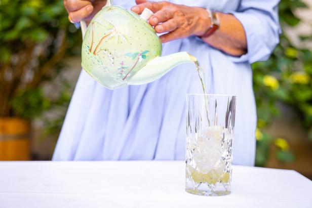 Eine Person gießt ein Getränk aus einer verzierten Teekanne in ein Glas mit Eiswürfeln.