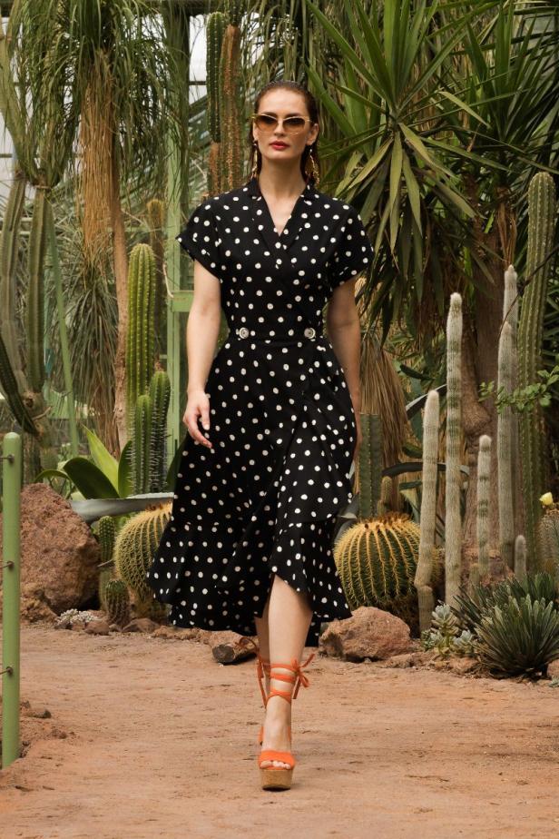 Eine Frau in einem gepunkteten Kleid und orangefarbenen Sandalen geht durch einen botanischen Garten.