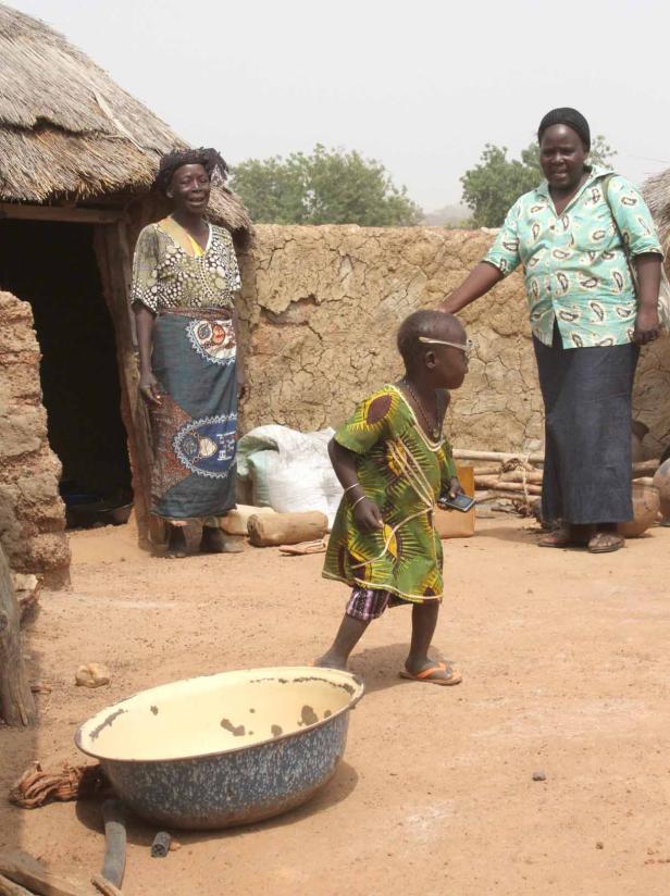 Drei Personen stehen vor einem Lehmhaus in einem afrikanischen Dorf.