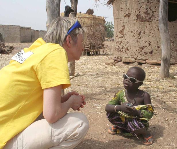 Eine Frau spricht mit einem Kind, das eine Sonnenbrille trägt und auf dem Boden sitzt.