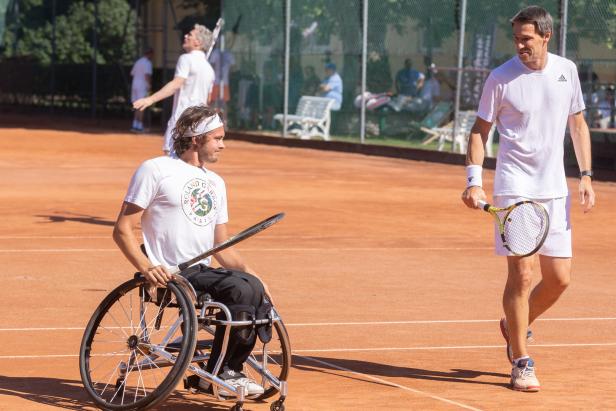 Ein Rollstuhltennisspieler auf einem Sandplatz mit einem stehenden Tennisspieler im Hintergrund.