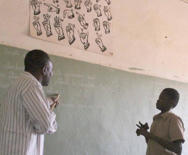 Ein Lehrer erklärt einem Schüler die Gebärdensprache vor einer Tafel.