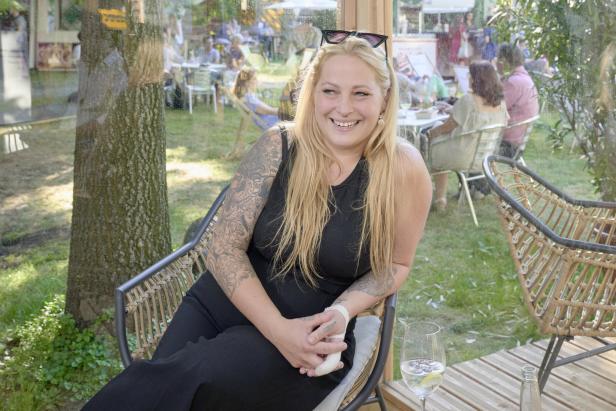 Eine lächelnde Frau mit Tätowierungen sitzt in einem Café im Freien.
