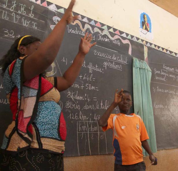 Eine Lehrerin und ein Schüler stehen vor einer Tafel mit Rechenaufgaben in einem Klassenzimmer.