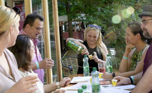Eine Gruppe von Menschen sitzt an einem Tisch, während eine Frau Getränke aus einer grünen Flasche einschenkt.