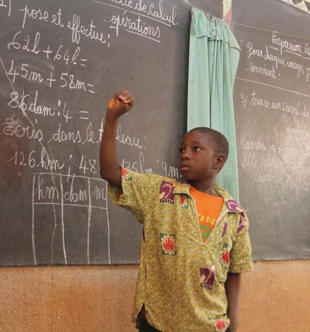 Ein Junge steht vor einer Tafel mit mathematischen Aufgaben und meldet sich.