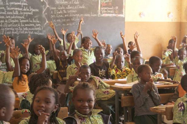 Eine Klasse von Kindern sitzt in einem Klassenzimmer und hebt die Hände.