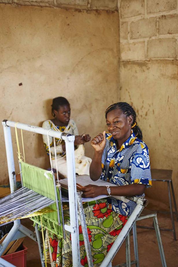 Eine Frau webt an einem Webstuhl, während eine andere Frau im Hintergrund sitzt.
