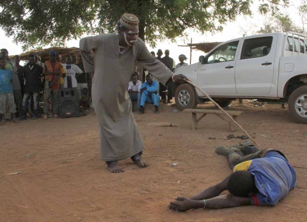 Ein Mann mit Gesichtsbemalung steht mit einem Stock über einer am Boden liegenden Person.