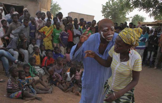 Ein Mann mit Gesichtsbemalung tanzt mit einer Frau vor einer Gruppe von Menschen in einem Dorf.
