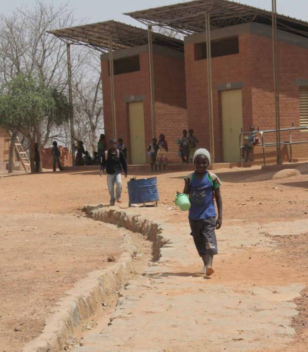 Ein Kind trägt einen Eimer auf einem sandigen Schulhof mit Ziegelgebäuden im Hintergrund.