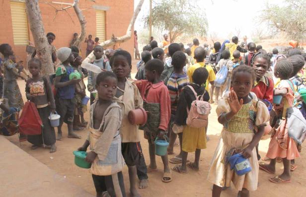 Eine Gruppe afrikanischer Kinder steht im Freien, viele tragen Behälter oder Taschen.