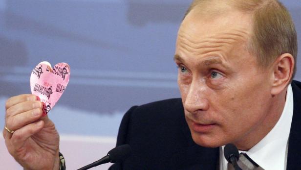 Wladimir Putin hält ein herzförmiges Objekt mit russischer Schrift in der Hand.