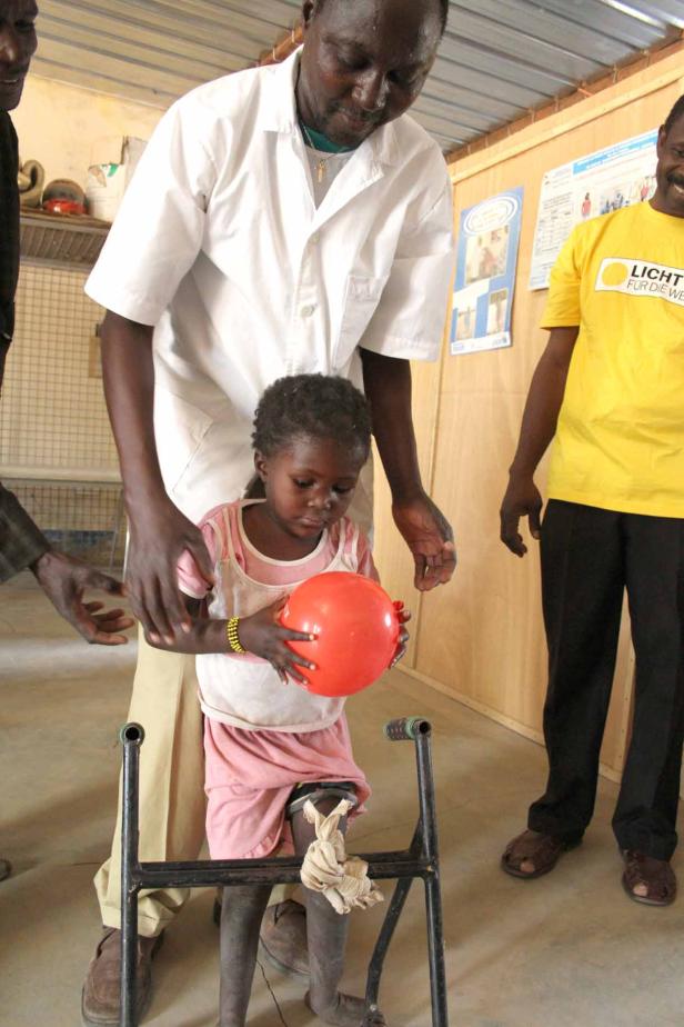 Ein Arzt hilft einem kleinen Mädchen mit Gehgestell, einen roten Ball zu halten.