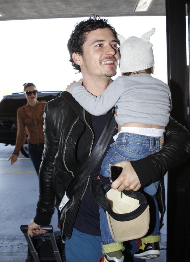 Orlando Bloom trägt sein Kind, während Miranda Kerr im Hintergrund zu sehen ist.