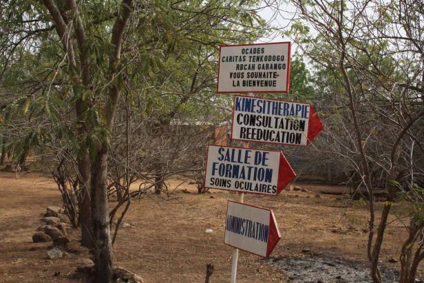 Mehrere Schilder weisen den Weg zu Kinesitherapie, Beratung, Umerziehung, Augenklinik und Verwaltung.
