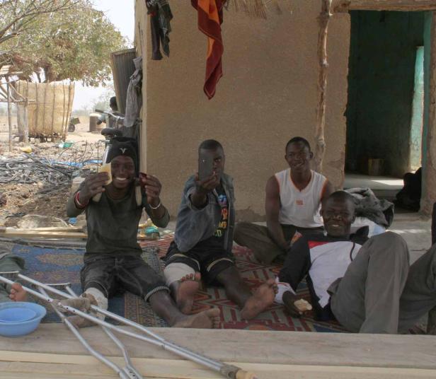 Vier Männer sitzen barfuß vor einem Haus, einer macht ein Foto mit seinem Handy.