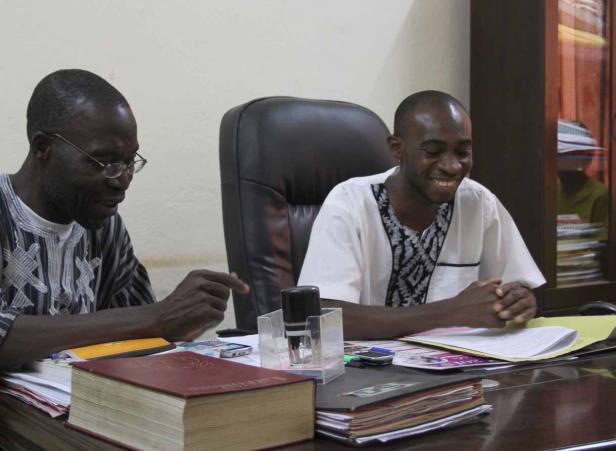 Zwei Männer sitzen an einem Schreibtisch mit Büchern und Dokumenten.