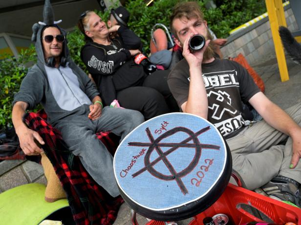 Eine Gruppe von Menschen sitzt im Freien, einer trinkt aus einer Flasche, ein anderer trägt ein Tierkostüm.