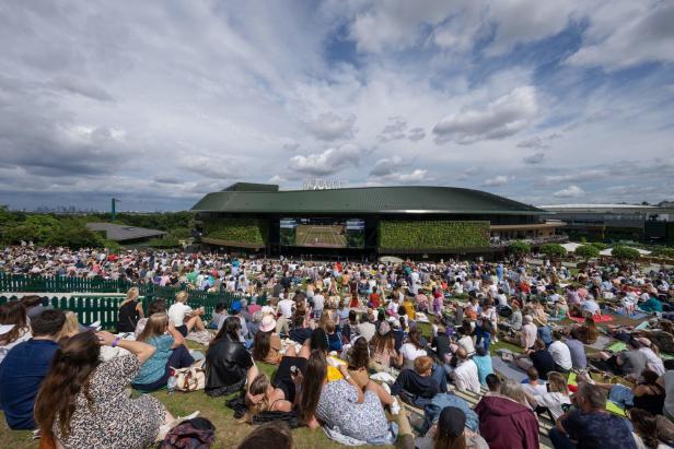 Viele Menschen sitzen auf dem Rasen vor einer großen Leinwand und schauen Tennis in Wimbledon.