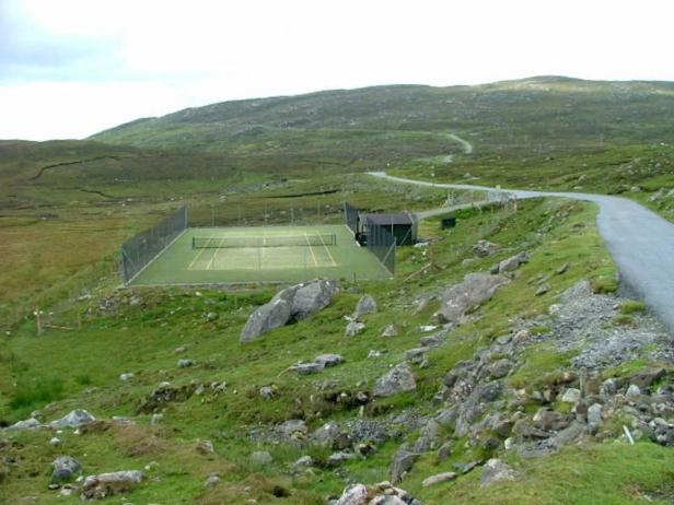 Ein Tennisplatz liegt in einer grünen, hügeligen Landschaft neben einer Straße.