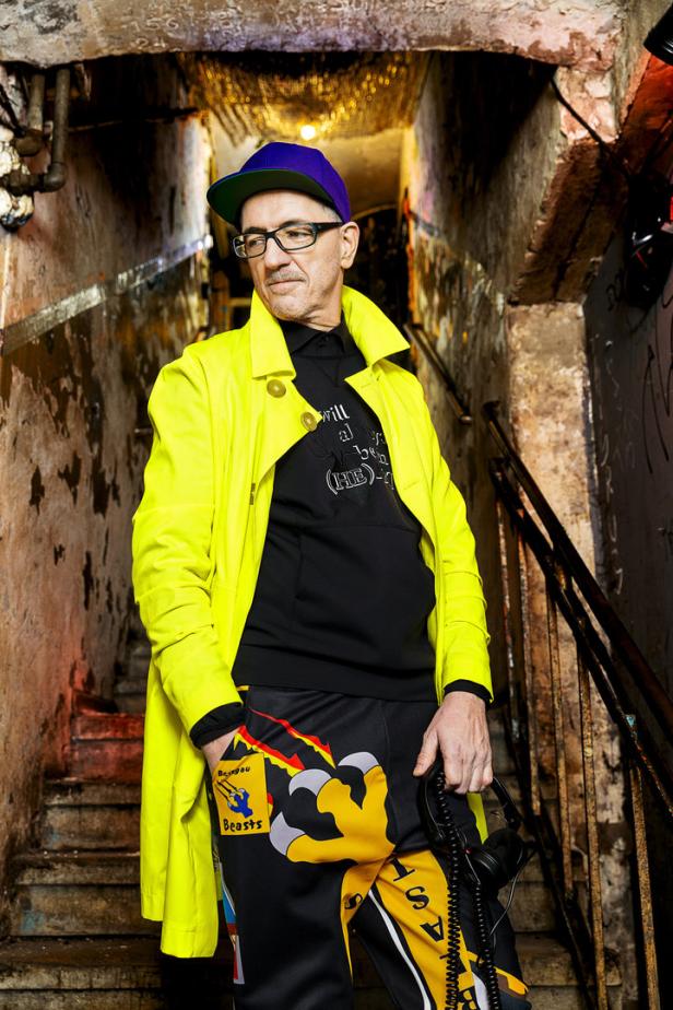 Ein Mann mit Brille, neonfarbenem Mantel und Kappe steht in einem Treppenhaus.