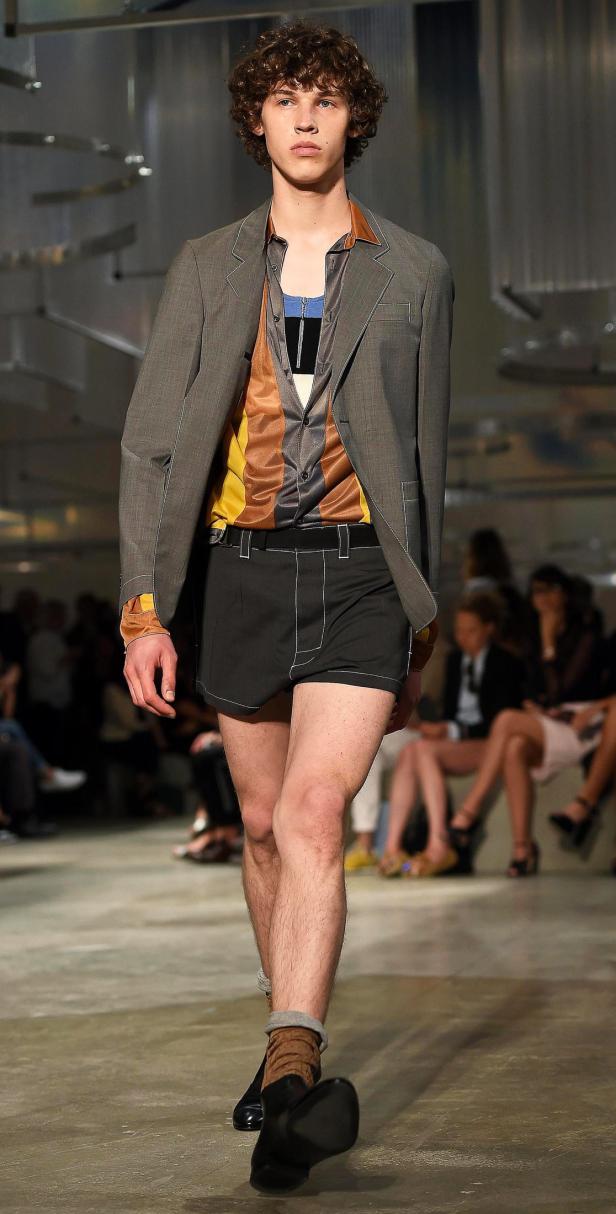 Ein Mann mit lockigem Haar trägt einen grauen Blazer und kurze, dunkle Hosen auf einem Laufsteg.