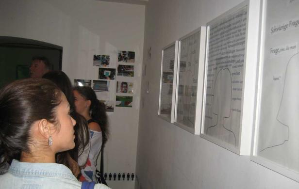 Besucher betrachten eine Ausstellung mit gerahmten Texten und Zeichnungen an einer Wand.